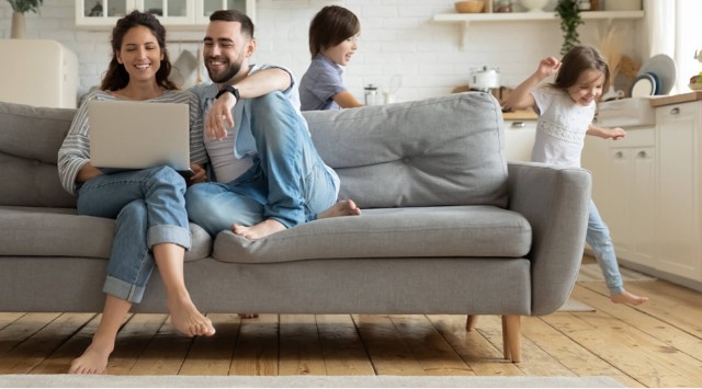 couple on sofa with laptop, children playing behind