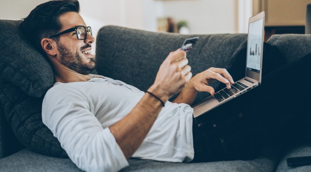 man in glasses laying on a sofa while online shopping with a laptop